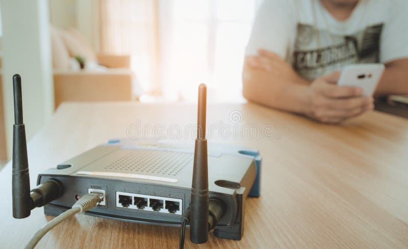 Primer de un router inalámbrico y de un hombre joven que usa un smartphone en sala de estar en casa con una ventana en el fondo imagen de archivo libre de regalías Primer de un router inalámbrico y de un hombre joven que usa un smartphone en sala de estar en casa con una ventana en el fondo imagen de archivo libre de regalías
