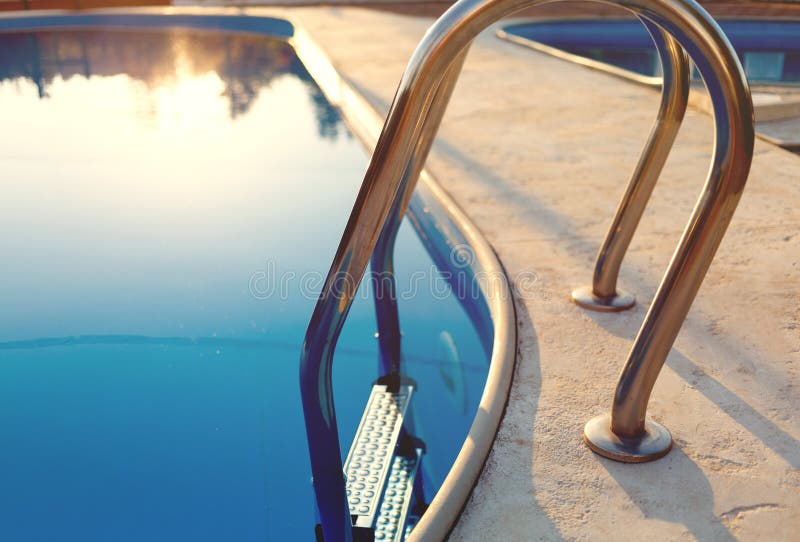 Primer de una parte de la piscina con una escalera de acero inoxidable y del agua azul en puesta del sol Las vacaciones de verano foto de archivo libre de regalías Primer de una parte de la piscina con una escalera de acero inoxidable y del agua azul en puesta del sol Las vacaciones de verano foto de archivo libre de regalías
