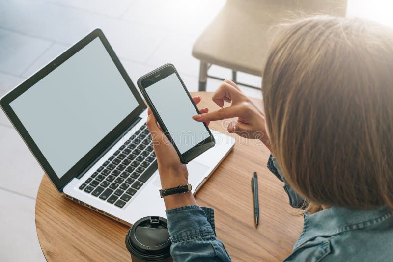 Primer del smartphone con la pantalla en blanco en las manos de la mujer joven que se sientan en la tabla de madera redonda y la imagen de archivo