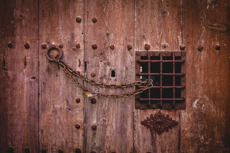 Primer tirado de una cerradura de cadena oxidada vieja en una puerta de madera grande con una pequeña cerca del metal foto de archivo Primer tirado de una cerradura de cadena oxidada vieja en una puerta de madera grande con una pequeña cerca del metal foto de archivo