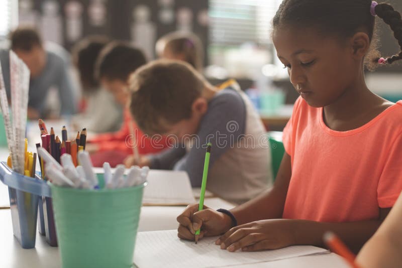 Primo piano di una ragazza razza mista della scuola che scrive sul suo taccuino in un'aula fotografia stock Primo piano di una ragazza razza mista della scuola che scrive sul suo taccuino in un'aula fotografia stock