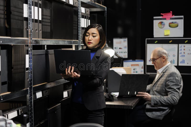 Asian private investigator in agency depository office room looking for criminal case file paperwork on cabinet shelves. Detective women and elderly assistant coworker surrounded by criminology folders. Asian private investigator in agency depository office room looking for criminal case file paperwork on cabinet shelves. Detective women and elderly assistant coworker surrounded by criminology folders