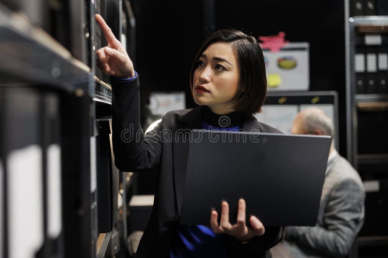 Asian private investigator in criminal cases archival depository office looking for criminal case paperwork on cabinet shelves. Detective surrounded by criminology file folders in agency cabinet room. Asian private investigator in criminal cases archival depository office looking for criminal case paperwork on cabinet shelves. Detective surrounded by criminology file folders in agency cabinet room