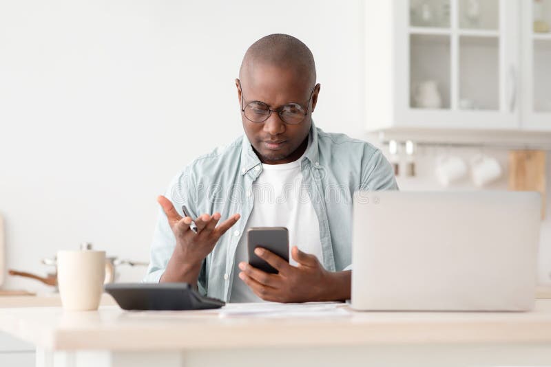 Problema financiero. hombre afroamericano enojado calculando cargos de servicios públicos y facturas usando calculadora y teléfono sentado en la cocina en casa. concepto de ahorro y finanzas