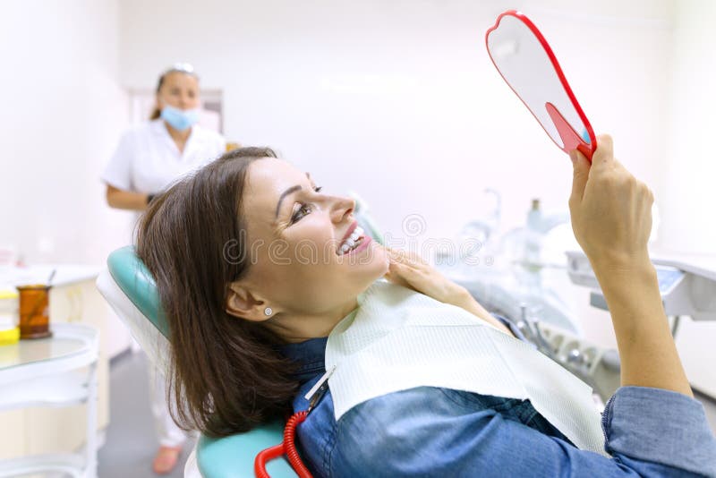 Proceso del tratamiento dental Paciente femenino que mira sus dientes en espejo mientras que se sienta en silla dental imagen de archivo Proceso del tratamiento dental Paciente femenino que mira sus dientes en espejo mientras que se sienta en silla dental imagen de archivo