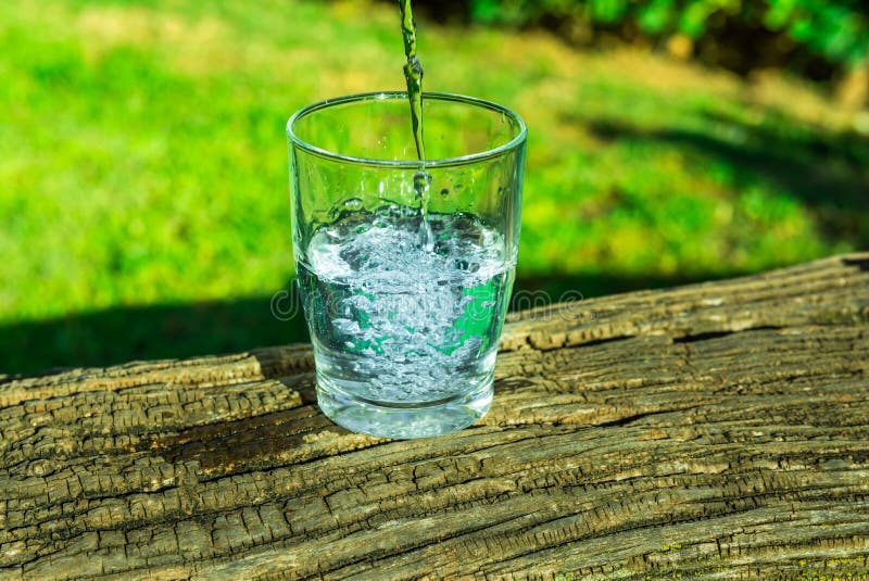 Process of pouring pure clear water into a glass from top, wooden log, green grass in the background, outdoors, health, hydration, cleansing, rejuvenation. Process of pouring pure clear water into a glass from top, wooden log, green grass in the background, outdoors, health, hydration, cleansing, rejuvenation