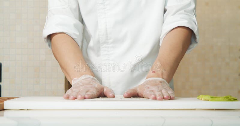 Proche des mains masculines de chef de file dans les gants déroulant le tapis en bambou pour faire des sushis dans la cuisine