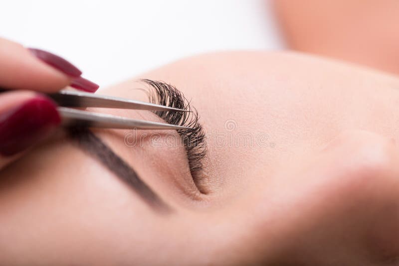 Close up cosmetician hand sticking artificial lash to female eye by tweezers. Close up cosmetician hand sticking artificial lash to female eye by tweezers