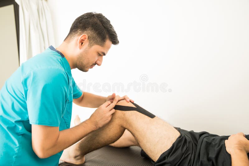Profesional de la fisioterapia que aplica la cinta del KT en rodilla del ` s del atleta fotografía de archivo libre de regalías Profesional de la fisioterapia que aplica la cinta del KT en rodilla del ` s del atleta fotografía de archivo libre de regalías
