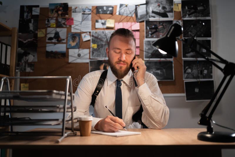 A professional detective works on a murder in the office at night. Sitting at the table takes a call from a witness and writes down the testimony in a notebook. copy space. A professional detective works on a murder in the office at night. Sitting at the table takes a call from a witness and writes down the testimony in a notebook. copy space.