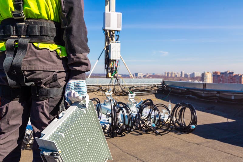 Professional industrial climber in uniform with telecomunication equipment in his hand and antennas of GSM DCS UMTS LTE bands, outdoor radio units on the roof. Working process of upgrading or swap telecommunication equipment. Professional industrial climber in uniform with telecomunication equipment in his hand and antennas of GSM DCS UMTS LTE bands, outdoor radio units on the roof. Working process of upgrading or swap telecommunication equipment