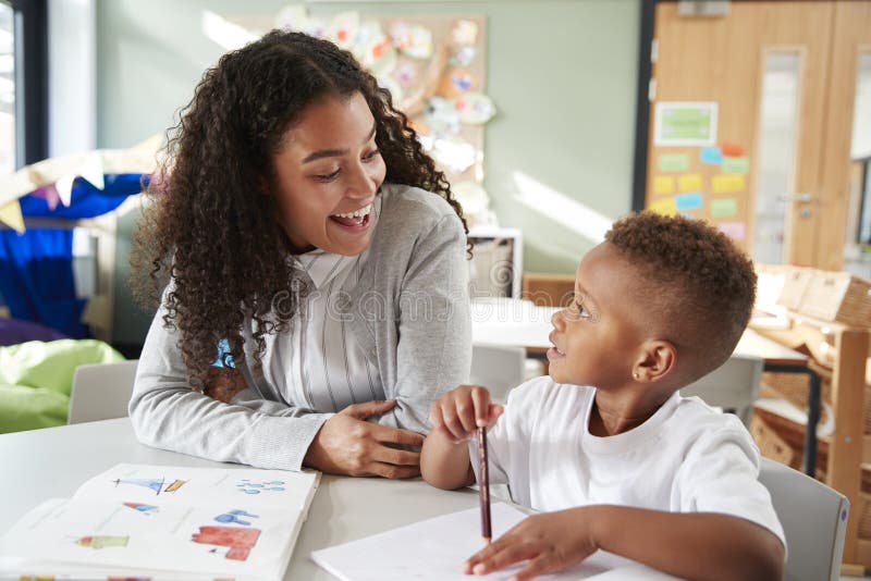 Female infant school teacher working one on one with a young schoolboy, sitting at a table smiling at each other, close up. Female infant school teacher working one on one with a young schoolboy, sitting at a table smiling at each other, close up