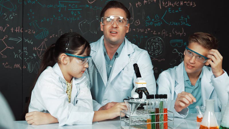 Professeur créatif pointant vers le tableau noir en parlant à un garçon intelligent. édification. Professeur créatif pointant la chimie au tableau noir tout en parlant à garçon du lycée à table avec microscope et tube de test avec liquide coloré. jeune étudiant portant un manteau de laboratoire en classe tige.