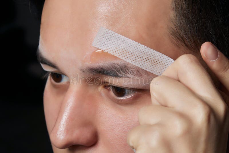 Professional barber doing threading procedure and correcting shape of eyebrows to young male client with tweezer in royalty free stock photo