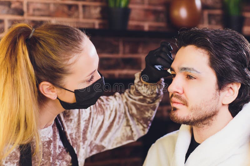 Professional barber in white shirt doing threading procedure and correcting shape of eyebrows to young male client stock images