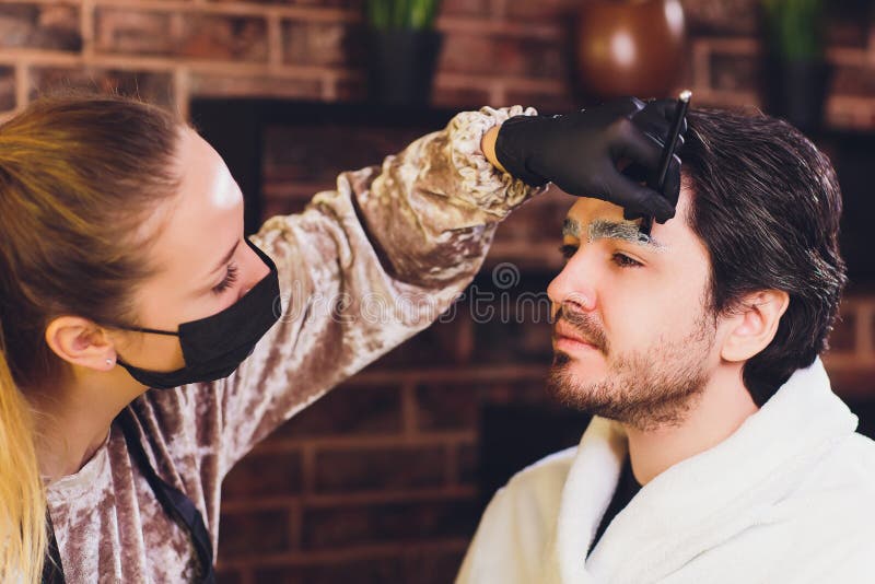 Professional barber in white shirt doing threading procedure and correcting shape of eyebrows to young male client stock images