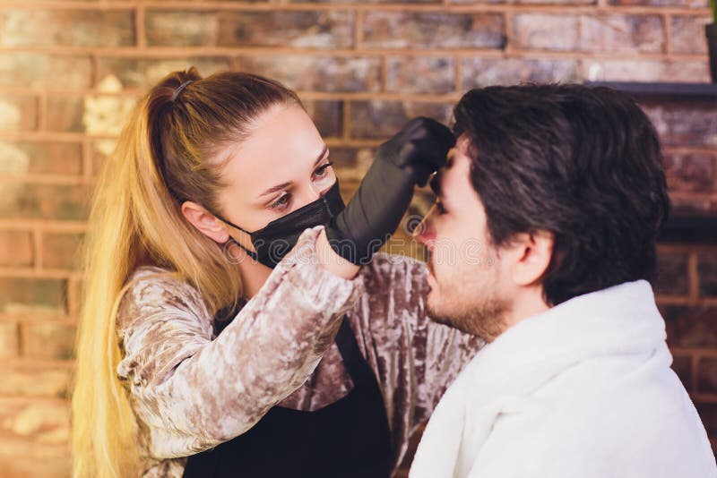 Professional barber in white shirt doing threading procedure and correcting shape of eyebrows to young male client royalty free stock images