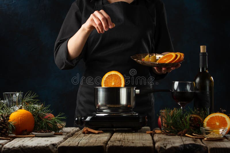 Professional chef pours orange into pan for preparing mulled wine on rustic wooden table with festive composition background.