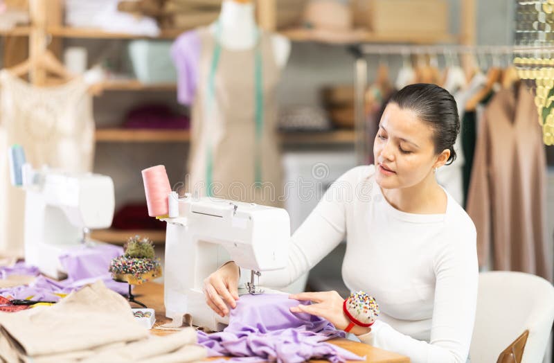 Professional young attentive woman seamstress sewing textile using sewing machine sitting at table at workplace in stock photo