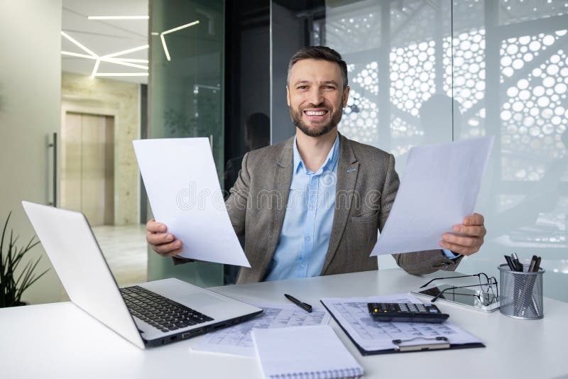 Smiling businessman sitting at a desk in a modern office, holding papers, with a laptop and calculator nearby. Portrays a productive and positive work environment. Smiling businessman sitting at a desk in a modern office, holding papers, with a laptop and calculator nearby. Portrays a productive and positive work environment.