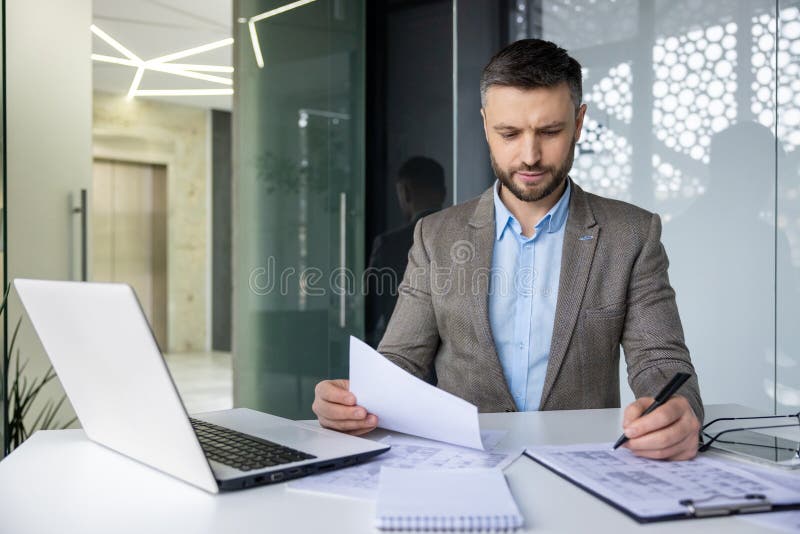 Focused businessman analyzing paperwork in a well-lit, contemporary office setting with a laptop and modern design elements. Focused businessman analyzing paperwork in a well-lit, contemporary office setting with a laptop and modern design elements.