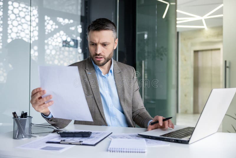 Focused business man reviewing paperwork while working on a laptop in a contemporary office setting, showing dedication and attention to detail. Focused business man reviewing paperwork while working on a laptop in a contemporary office setting, showing dedication and attention to detail.