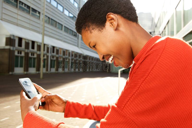 Profil de la jeune femme de couleur heureuse s'asseyant dans la ville et regardant le téléphone portable images libres de droits Profil de la jeune femme de couleur heureuse s'asseyant dans la ville et regardant le téléphone portable images libres de droits