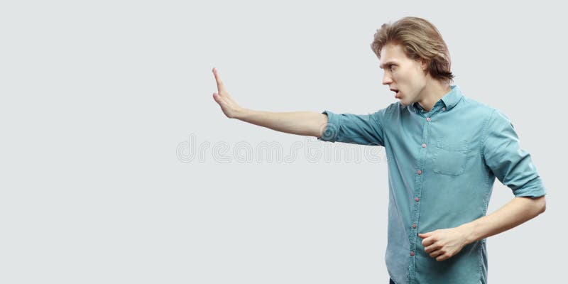 Profile side view portrait of serious handsome long haired blonde young man in blue casual shirt standing with stop hand gesture