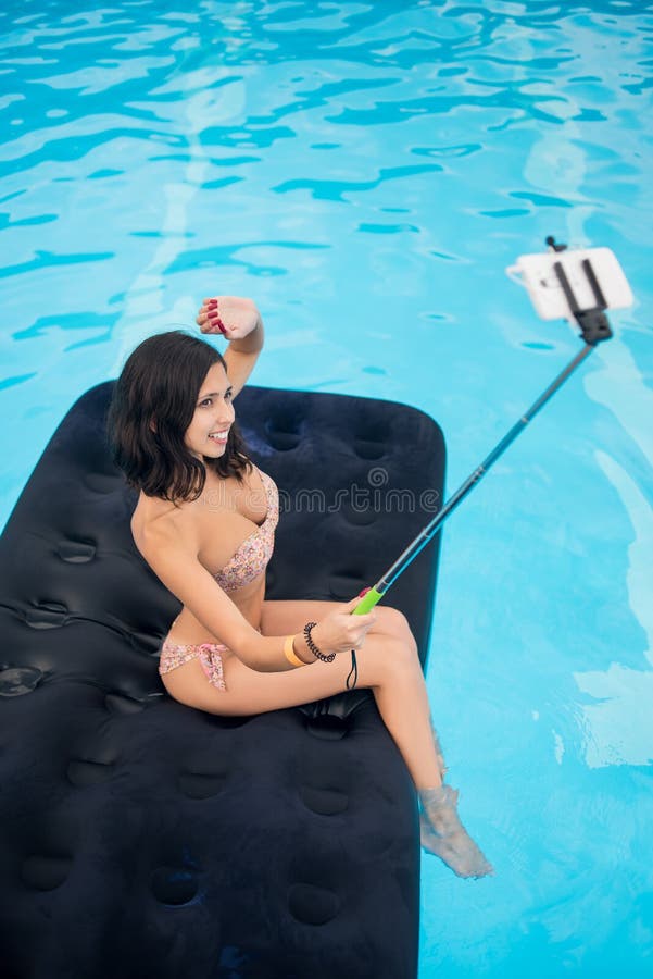 Profile of a young brunette makes selfie photo on the phone with selfie stick on mattress in pool. Copy space. View from above