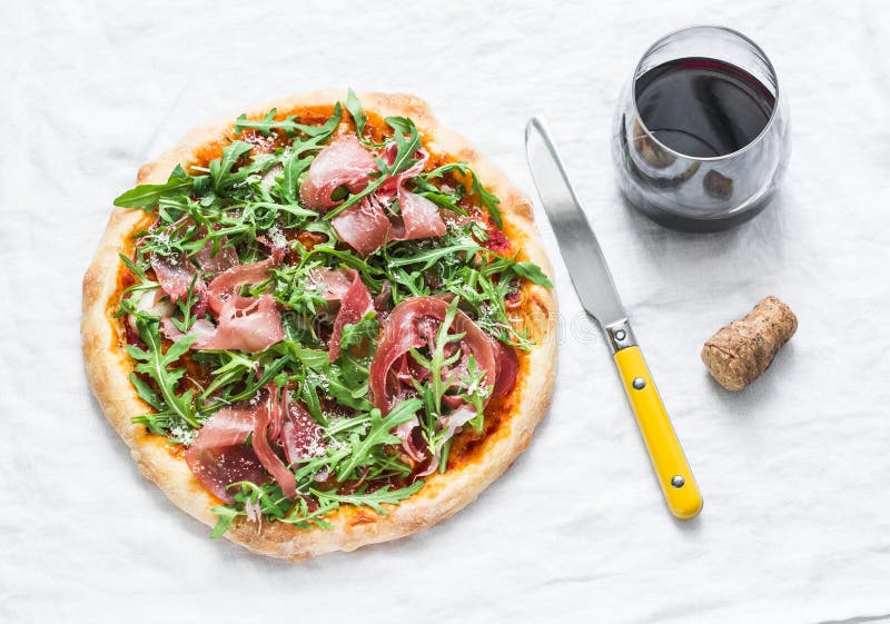 Prosciutto arugula homemade pizza and a glass of red wine on light background, top view. Delicious appetizers royalty free stock photography Prosciutto arugula homemade pizza and a glass of red wine on light background, top view. Delicious appetizers royalty free stock photography