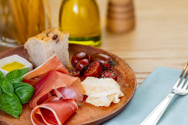 Italian ham, parmesan cheese, focaccia, tomato and basil twig in a wooden plate by a pasta jar, an olive oil bottle and cutlery stock photo Italian ham, parmesan cheese, focaccia, tomato and basil twig in a wooden plate by a pasta jar, an olive oil bottle and cutlery stock photo