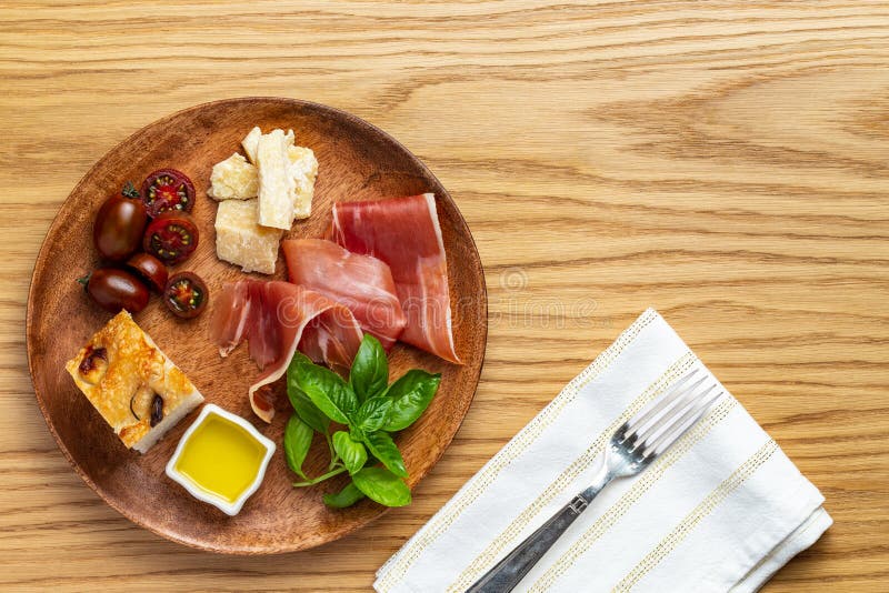Italian ham, parmesan cheese, focaccia, tomato and basil twig in a wooden plate by a white napkin on a wood background copy space royalty free stock images Italian ham, parmesan cheese, focaccia, tomato and basil twig in a wooden plate by a white napkin on a wood background copy space royalty free stock images