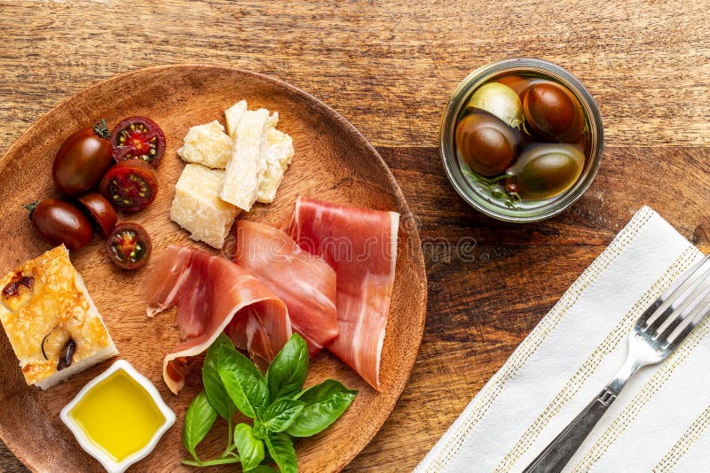 Italian ham, parmesan cheese, focaccia, tomato and basil twig in a wooden plate by a white napkin on a chopping board background. stock image Italian ham, parmesan cheese, focaccia, tomato and basil twig in a wooden plate by a white napkin on a chopping board background. stock image