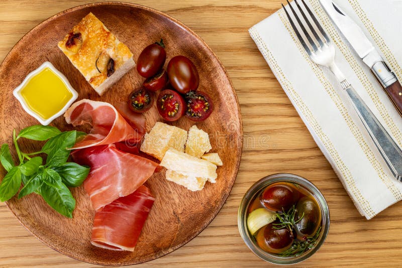 Italian ham, parmesan cheese, focaccia, tomato and basil twig in a wooden plate by a white napkin with cutlery on oak royalty free stock photography Italian ham, parmesan cheese, focaccia, tomato and basil twig in a wooden plate by a white napkin with cutlery on oak royalty free stock photography