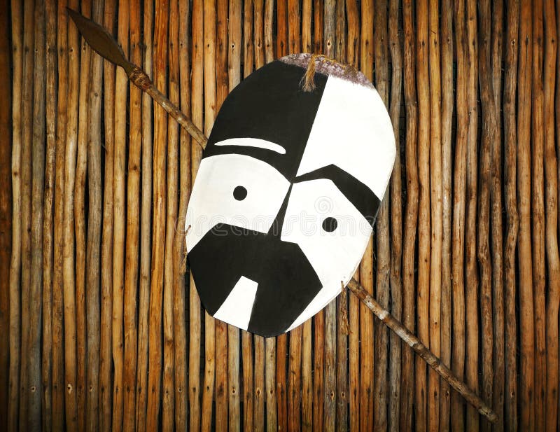 A photograph of a beautiful hand crafted tribal shield and spear from the Africa zulu warriors tribe. Pattern of black and white zulu mask on shield. Taken with background of wooden hut wall. Beautiful traditional African ethnic tribal art. A photograph of a beautiful hand crafted tribal shield and spear from the Africa zulu warriors tribe. Pattern of black and white zulu mask on shield. Taken with background of wooden hut wall. Beautiful traditional African ethnic tribal art.