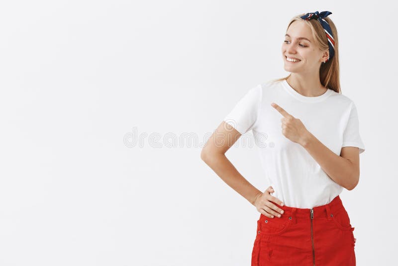 Proud mom showing child funny cloud on sky. Portrait of attractive happy and carefree blonde female in headband and red skrit, looking left and pointing at upper corner, smiling joyfully.