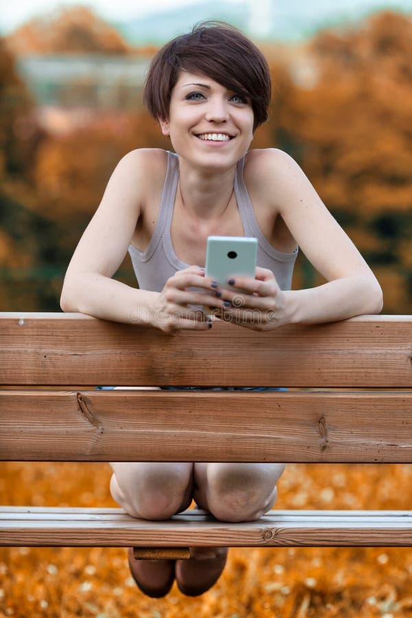 Friendly young woman smiling at the camera outdoors as she relaxes kneeling on a park bench holding her mobile phone. Friendly young woman smiling at the camera outdoors as she relaxes kneeling on a park bench holding her mobile phone