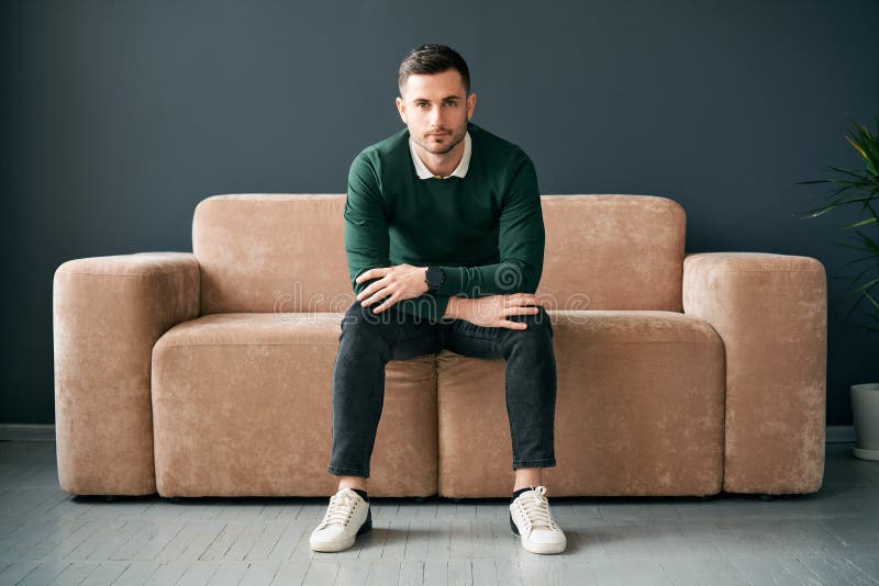 Przystojny modny mężczyzna pozujący się patrząc na kamerę siedząc na wygodnej sofie w nowoczesnym mieszkaniu