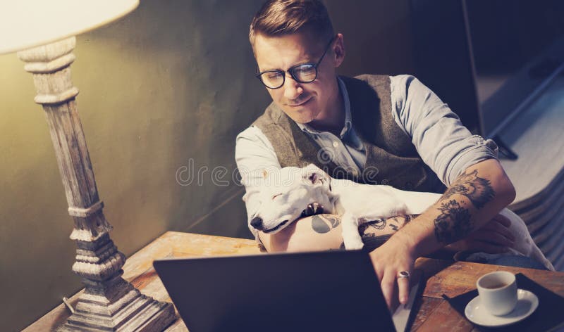 Przystojny tatuujący mężczyzna w eyeglasses pracuje w domu na laptopie podczas gdy siedzący przy drewnianym stołem z ślicznym psi fotografia royalty free Przystojny tatuujący mężczyzna w eyeglasses pracuje w domu na laptopie podczas gdy siedzący przy drewnianym stołem z ślicznym psi fotografia royalty free