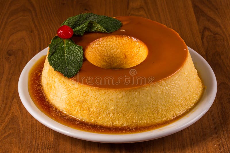 Pudim de leite, a delicious Brazilian flan dessert, with milk and condensed milk, topped with caramel sauce. It`s type of vanilla pudding. Served on white plate on wooden table.