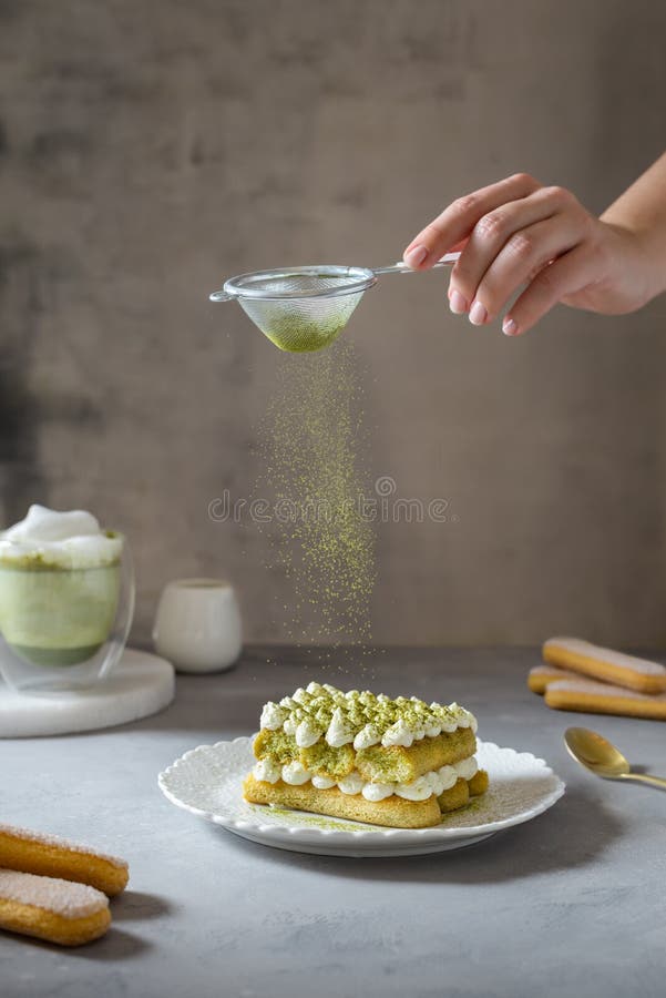 Female hand sprinkling matcha powder over tiramisu cake with green tea or spirulina. Superfoods, healthy nutrition. Confectionery, bakery, cafe menu concept. Side view. Female hand sprinkling matcha powder over tiramisu cake with green tea or spirulina. Superfoods, healthy nutrition. Confectionery, bakery, cafe menu concept. Side view