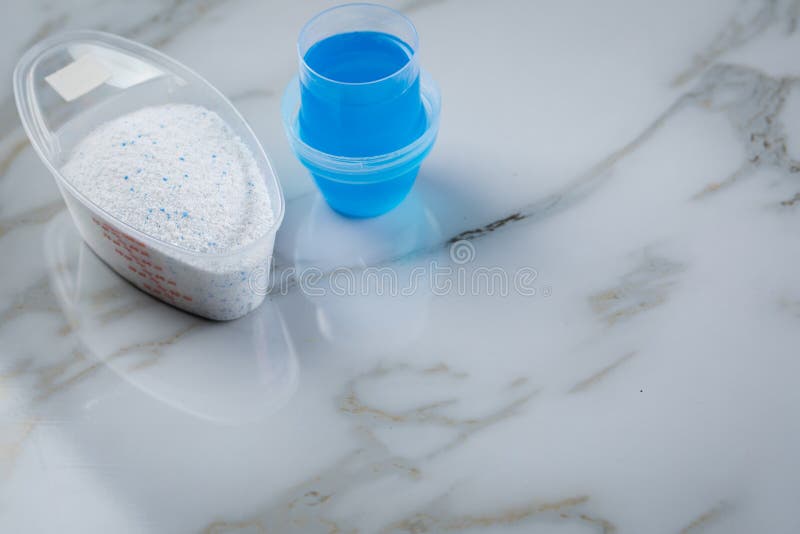 Laundry detergent powder and blue liquid gel in measuring cup on marble background