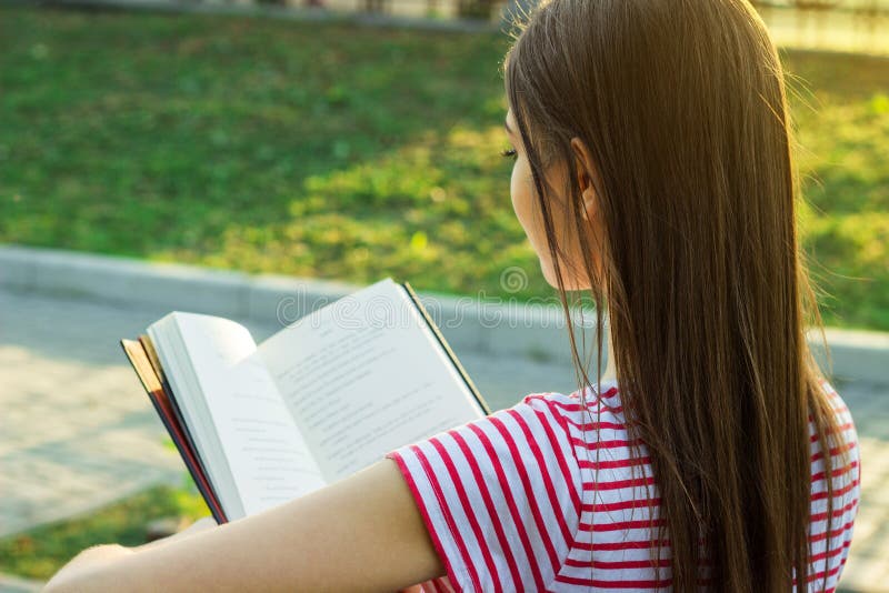 Punto di vista posteriore della ragazza adorabile con bei capelli lunghi che legge un libro fuori fotografia stock Punto di vista posteriore della ragazza adorabile con bei capelli lunghi che legge un libro fuori fotografia stock