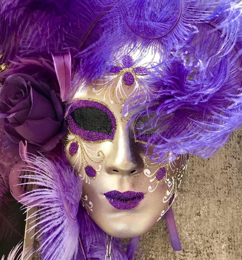 A purple and gold masquerade mask hanging on a wall in Venice Italy. The mask comprises glitter decoration, feathers and a rose. A good example of a traditional symbol of masquerade balls, theatre, costumes. A purple and gold masquerade mask hanging on a wall in Venice Italy. The mask comprises glitter decoration, feathers and a rose. A good example of a traditional symbol of masquerade balls, theatre, costumes