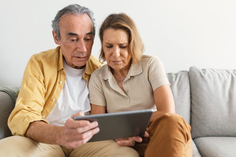 Puzzled senior spouses using digital tablet having hard time learning or browsing internet, sitting on sofa, copy space stock photos