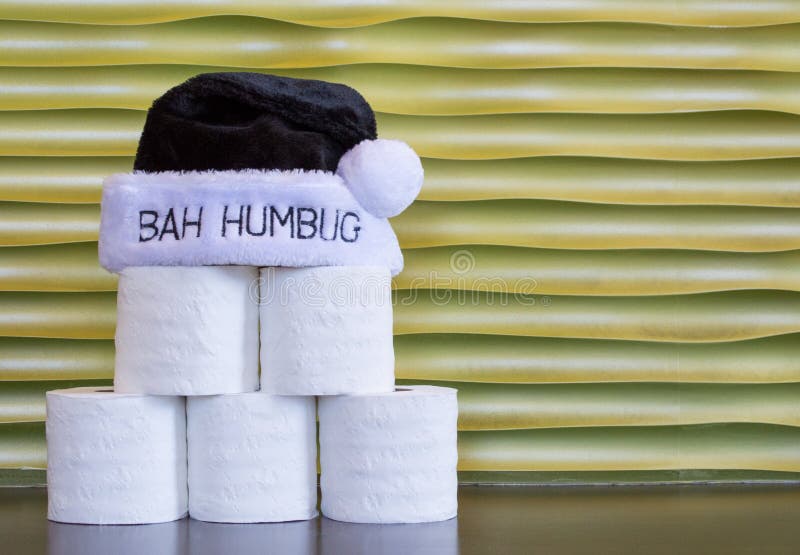 A pyramid of six rolls of toilet paper with a black fur Bah Humbug hat against a gold wall with copy space. A pyramid of six rolls of toilet paper with a black fur Bah Humbug hat against a gold wall with copy space