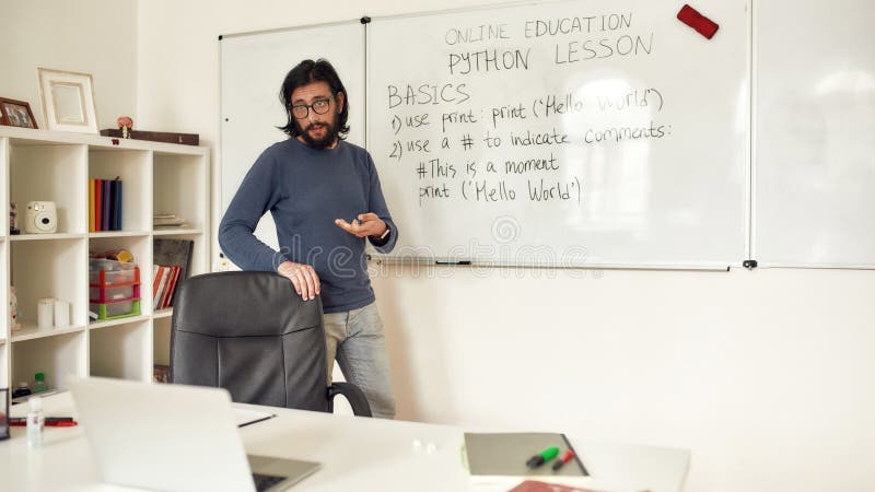 在线学习Python。年轻的白人男性教师戴着眼镜站在白板旁，进行Python在线课程 免版税库存图片