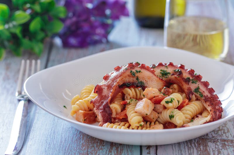 A plate of pasta with smoked swordfish, octopus and cherry tomatoes. In the background a glass of white wine, some green plants and purple hydrangea flowers. A plate of pasta with smoked swordfish, octopus and cherry tomatoes. In the background a glass of white wine, some green plants and purple hydrangea flowers