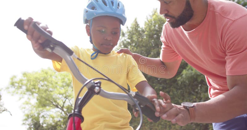 Père américain africain enseignant à son fils de faire du vélo dans un jardin ensoleillé ralenti banque de vidéos