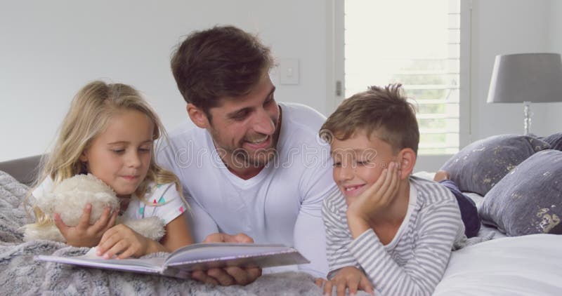 Père avec ses enfants lisant le livre d'histoire dans la chambre à coucher à la maison 4k